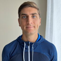 En mann med kort hår, Mirko Vicentijevic, i en blå atletisk jakke smiler subtilt mot et innendørs bakteppe med en lys gardin til høyre.