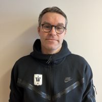 Magnus Olsen, iført briller og svart Nike-hettegenser med hvit logo, smiler lett til kameraet i en nøytral innendørs setting.