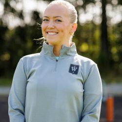 Et portrett av Kristine Årvik, en atletisk kvinne som smiler selvsikkert utendørs, kledd i en sporty grå genser, klar for en treningsøkt eller utendørsaktivitet.