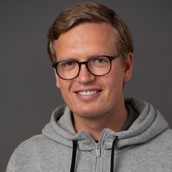 En smilende mann med blondt hår iført briller og grå hettegenser mot grå bakgrunn er Andreas Gulbrandsen.