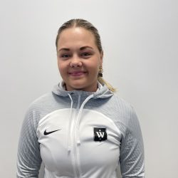 En person som har på seg en grå Nike-sportsjakke med en "W"-logo, identifisert som Mia Michelle Malmer, smiler til kameraet mot en vanlig bakgrunn.