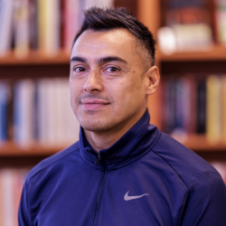 Elio Figueroa, en selvsikker mann med et lite smil, iført Nike-sportsjakke, står foran en bokhylle fylt med bøker.