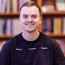En smilende Joakim Halvorsen i svart genser står foran en bokhylle fylt med bøker, og utstråler en vennlig og imøtekommende væremåte.