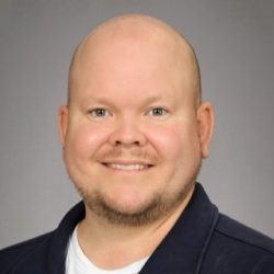 Portrett av en smilende mann med barbert hode og svart skjorte mot en grå bakgrunn.