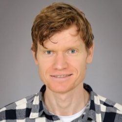 En vennlig utseende mann med rotete krøllete hår som smiler til kameraet, iført en rutete skjorte, mot en nøytral grå bakgrunn.