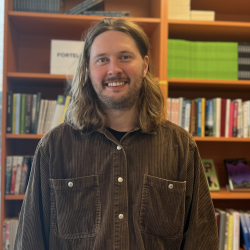 En smilende mann med langt hår poserer foran en bokhylle i et bibliotek eller en bokhandel.