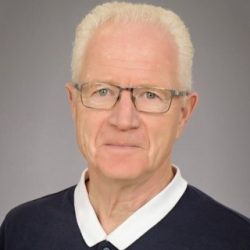 Portrett av en senior mann med hvitt hår, iført briller og en skjorte med blå krage, mot en grå bakgrunn.