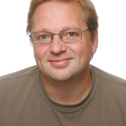 En smilende mann med lyst hår iført briller og en nøytralfarget skjorte mot en vanlig bakgrunn.