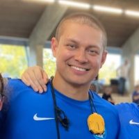 En smilende idrettsutøver iført en blå nike-skjorte med en gullmedalje rundt halsen og deler et øyeblikk av kameratskap.