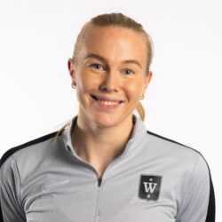 En person med lyst hår trukket tilbake smiler, iført en lysegrå atletisk jakke med glidelås med en svart stripe på ermene og et skjoldemblem med "W" på venstre bryst. Bakgrunnen er ensfarget hvit, noe som øker fokuset på Oda Cathrine Lunne Mastads muntre uttrykk.